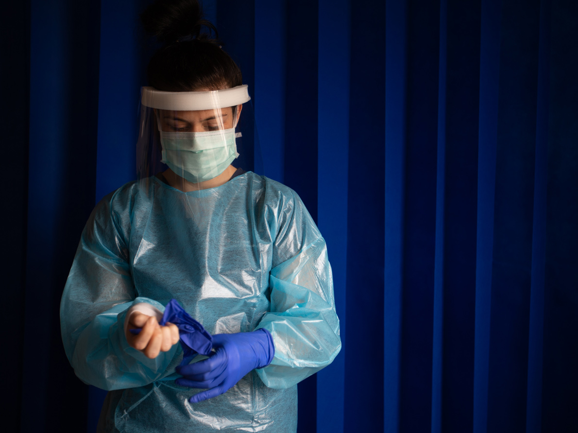 Image of a nurse in scrubs and face protection putting on protective gloves.